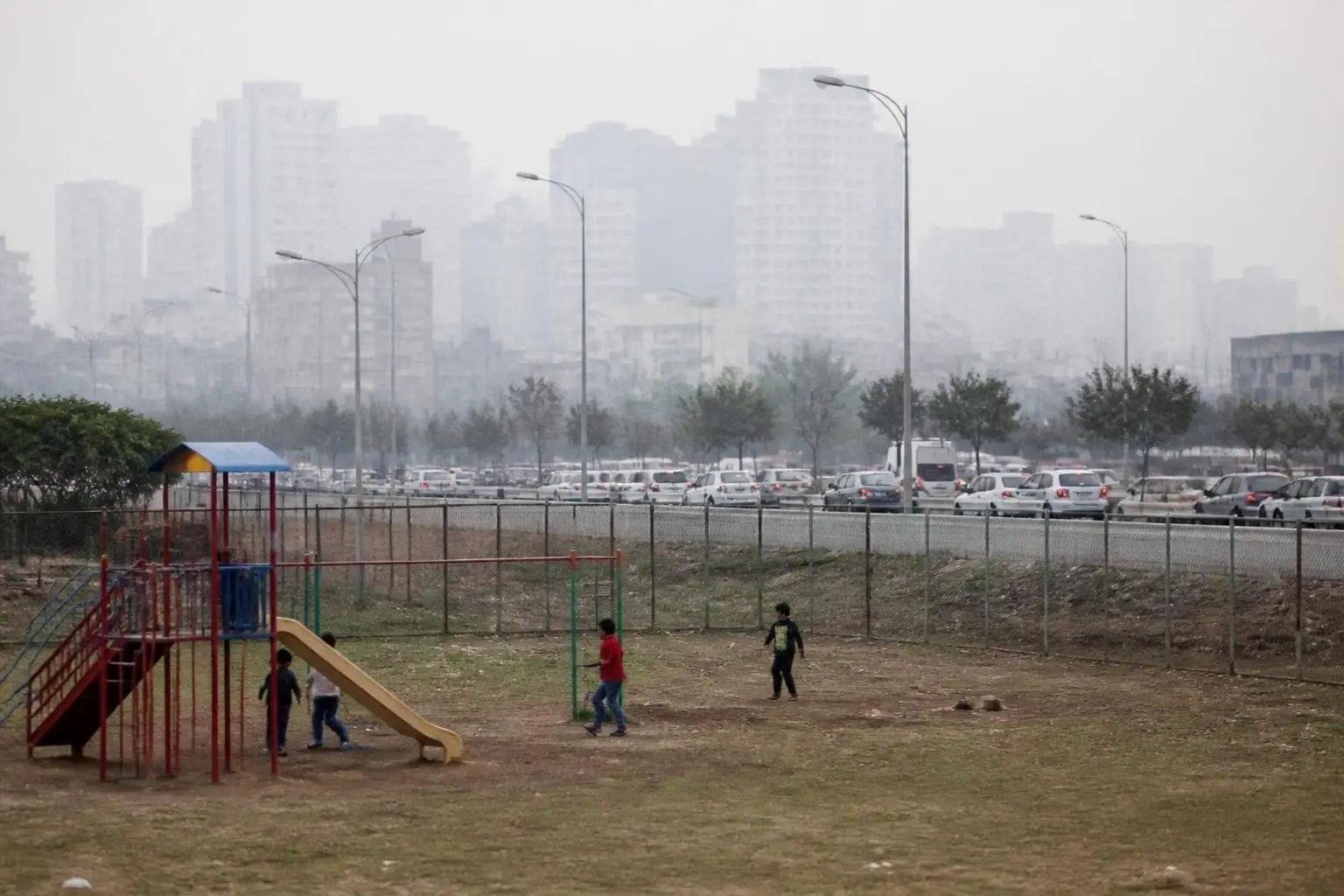 Young children seen from a distance playing on a city playground next to a road with passing traffic and light haze in the air