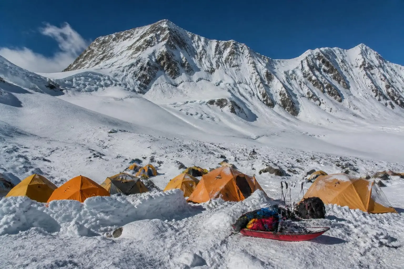 Cluster of yellow and orange expedition tents with snow walls and sleds on a glacier camp, a large snowy ridge behind under a clear sky
