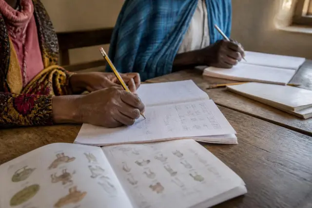 Adult hands holding a pencil over open exercise books with handwriting practice and a reading primer on a worn wooden desk in daylight