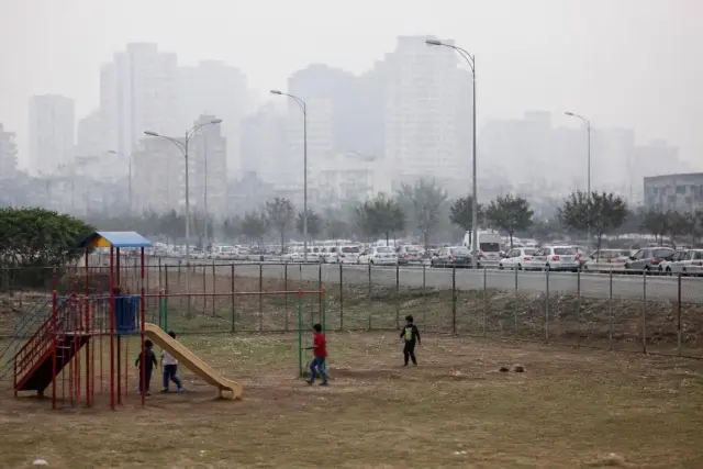 Young children seen from a distance playing on a city playground next to a road with passing traffic and light haze in the air