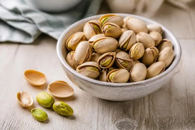 A small ceramic bowl of roasted pistachios in split shells on a light wooden table, with green kernels and empty shells beside it