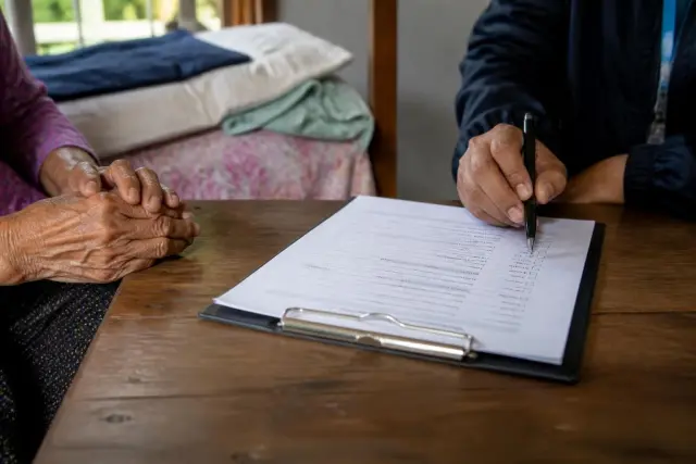 Hands of a fieldworker and an older adult at a wooden table with a clipboard questionnaire and a pen in warm window light